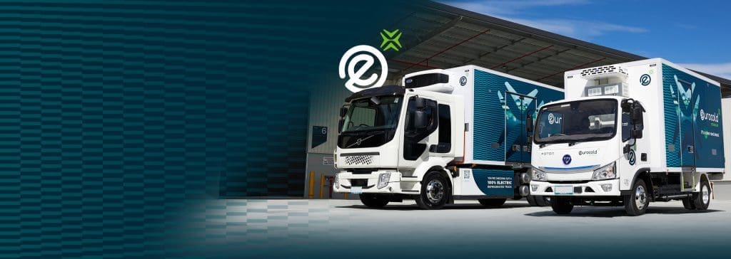 Two electric delivery trucks with blue and white branding are parked outside a modern warehouse under a blue sky, with company logos and eco-friendly symbols visible on the vehicles.